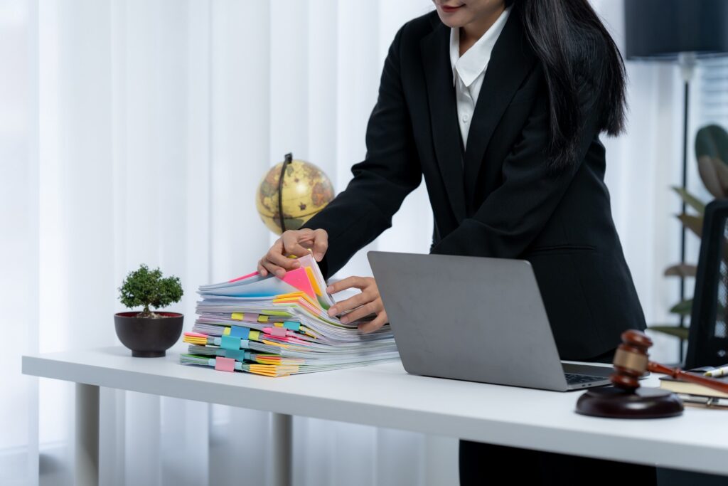 Professional woman organizing a large stack of documents with colorful tabs at a desk, with a laptop and gavel nearby.