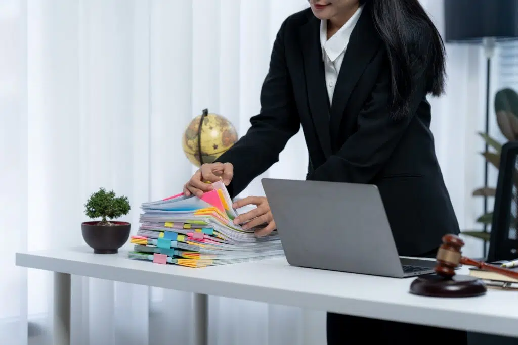 Professional woman organizing a large stack of documents with colorful tabs at a desk, with a laptop and gavel nearby.
