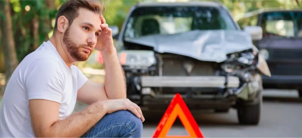 A man sitting on the road near a caution triangle in front of a car that has been in a car accident