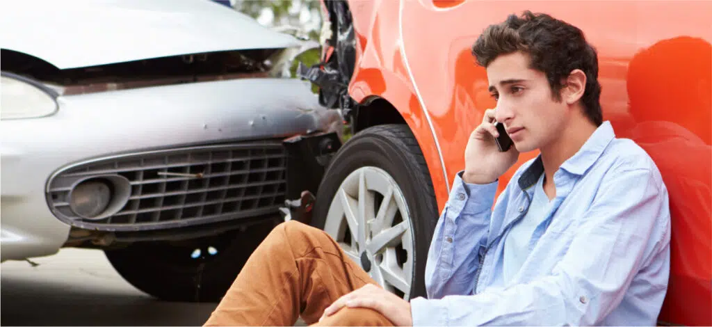 A man sitting on the ground next to his car. He is talking on a cell phone. after an accident