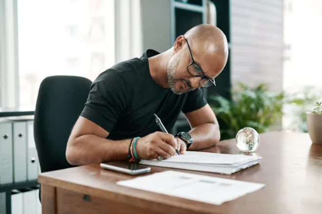 A man sitting at a desk, wearing glasses and writing on documents with focused attention, surrounded by paperwork and office supplies in a modern workspace.