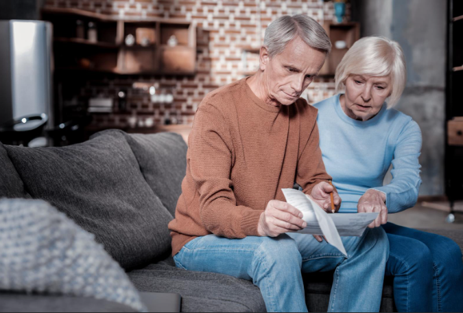 An older couple sitting on a couch, closely reviewing and discussing paperwork with serious expressions, suggesting concern over financial or legal documents.