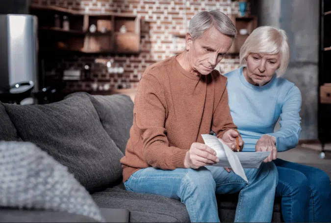 An older couple sitting on a couch, closely reviewing and discussing paperwork with serious expressions, suggesting concern over financial or legal documents.
