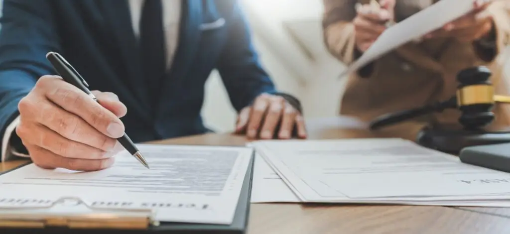 Closeup of paperwork on a desk and people in the background pointing to parts of it with a pen