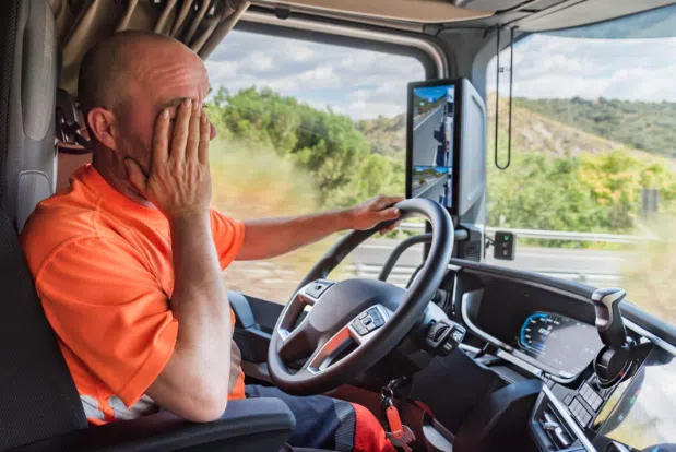 A tired truck driver in an orange shirt sitting in the cab of a truck, resting his face in his hand while driving on a highway.