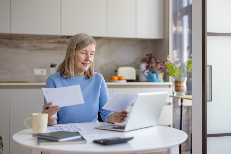 Woman reviewing paperwork while sitting at a table with a laptop in a bright kitchen.