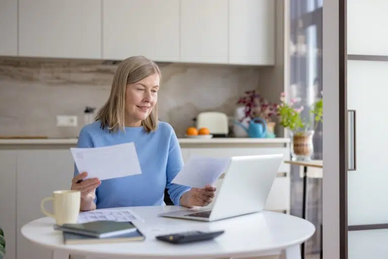 Woman reviewing paperwork while sitting at a table with a laptop in a bright kitchen.