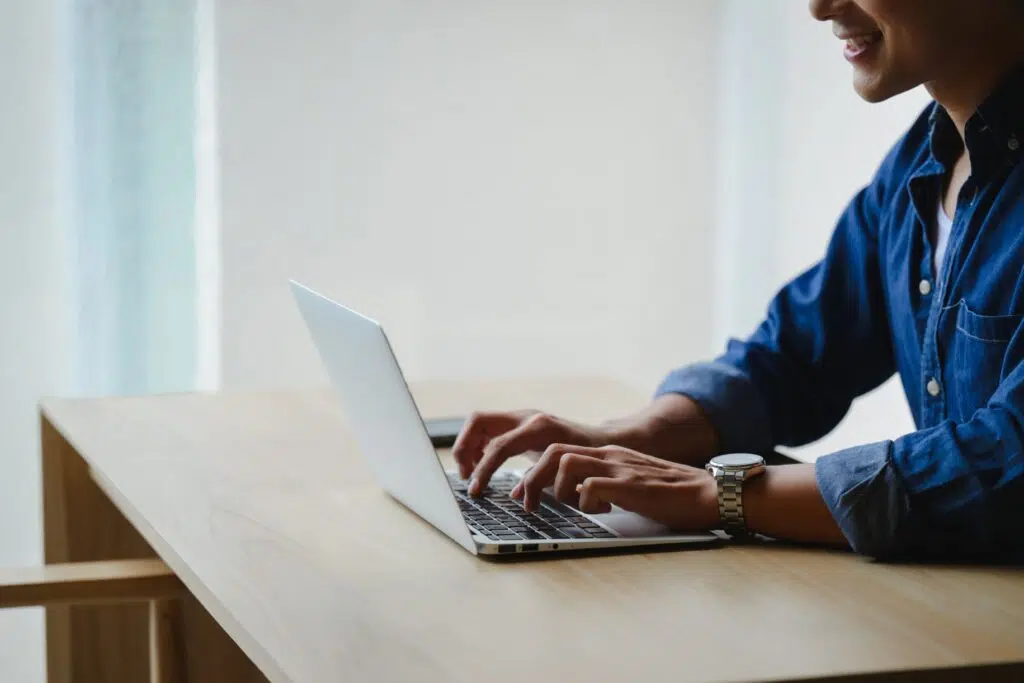 Person typing on a laptop while working at a wooden desk near a window.