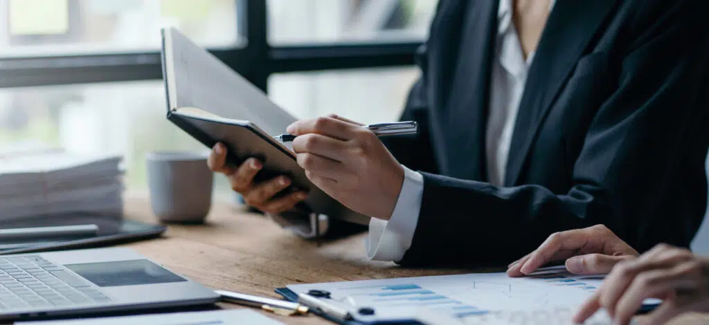 Closeup of two people's hands. One is pointing to notes in a book, and the other is looking at paperwork on a desk.