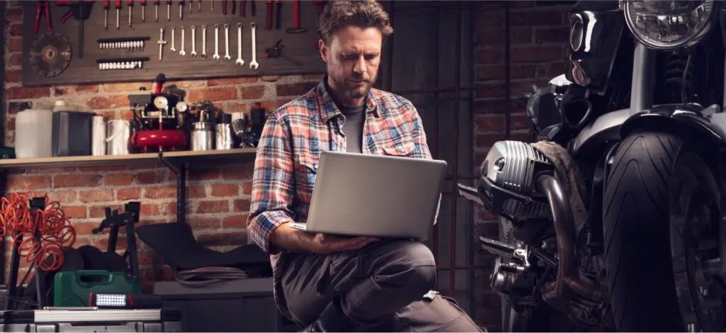 A person in a garage using a computer and sitting next to a motorcycle