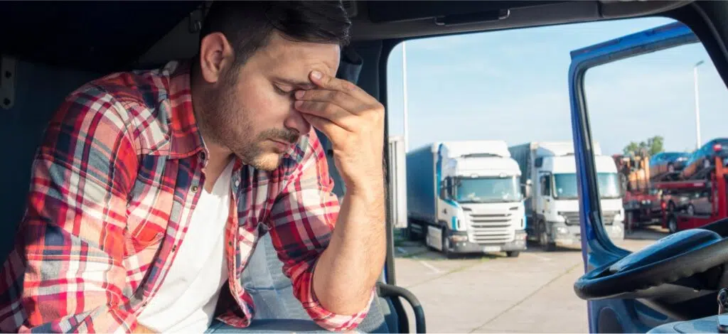 A truck driver sits in his cab, door open, holding his face due to fatigue.