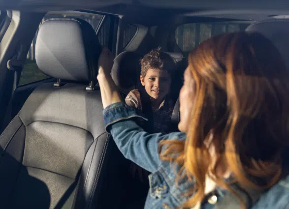 A woman in the front seat of a car turns around to talk to a smiling young boy sitting in the back seat. Sunlight streams in through the windows, illuminating both faces warmly.