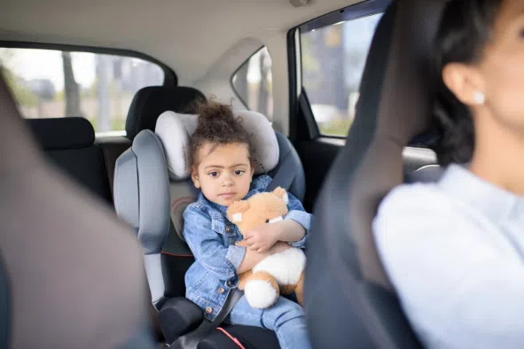A young girl sitting in a car seat in the back seat of a vehicle, holding a stuffed animal and looking out calmly. She is dressed in a denim outfit, while an adult woman drives the car.
