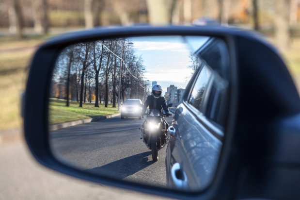 View of a motorcycle rider wearing a helmet reflected in a car’s side mirror, riding closely behind the vehicle on a city street.