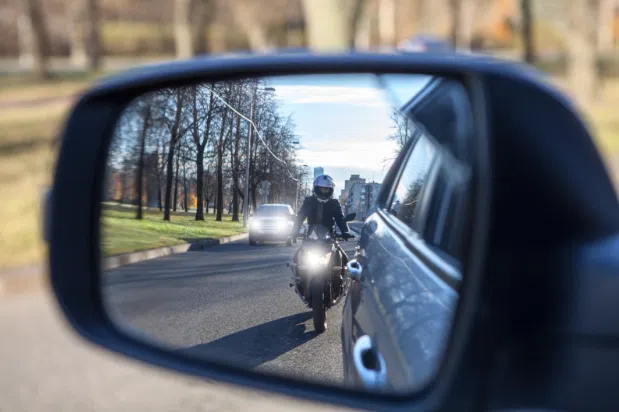 View of a motorcycle rider wearing a helmet reflected in a car’s side mirror, riding closely behind the vehicle on a city street.