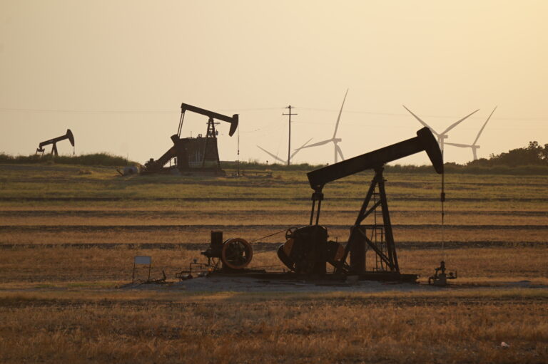 Oil pump jacks operating in an open field at sunset with wind turbines visible in the background, highlighting a contrast between traditional fossil fuel extraction and renewable energy sources.