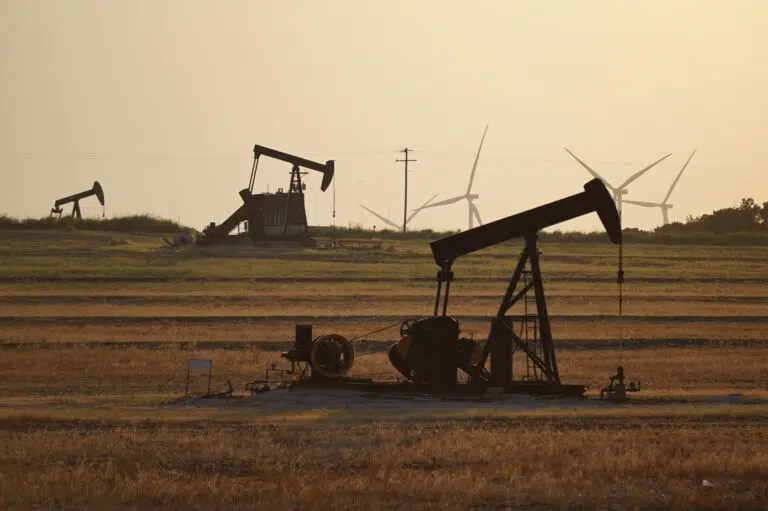 Oil pump jacks operating in an open field at sunset with wind turbines visible in the background, highlighting a contrast between traditional fossil fuel extraction and renewable energy sources.