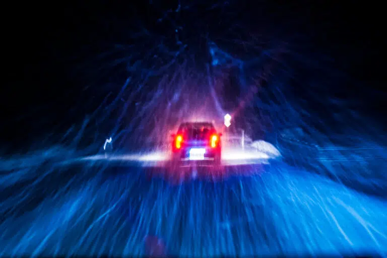 Car driving through heavy snow at night with headlights illuminating the road, symbolizing dangerous winter driving conditions or auto accidents in bad weather.
