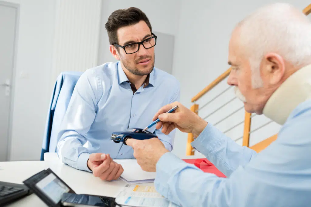 An injured older man wearing a neck brace discusses his car accident claim with an insurance adjuster or attorney, using a model car to explain the collision details during a consultation.