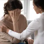 A supportive scene where one woman comforts another who is covering her face with her hand in sadness or emotional distress.