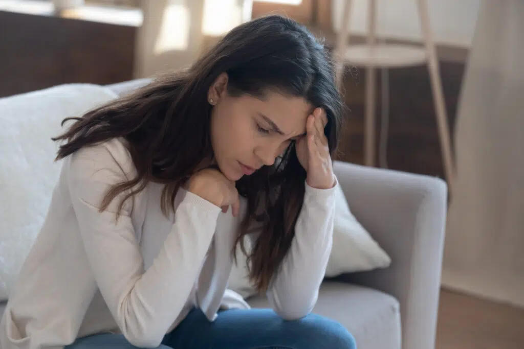 Unhappy young woman touching forehead, yearning, thinking about relationship problems, feeling depressed and lonely, sitting on couch alone, frustrated female suffering from headache or migraine