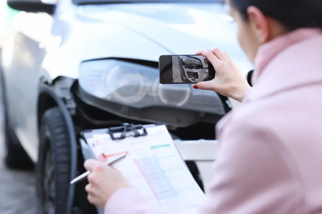 A woman documents car accident damage by taking a photo of a dented vehicle while holding an insurance claim form on a clipboard, representing car accident evidence collection or insurance inspection.