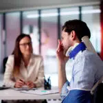 A man wearing a neck brace and arm sling sits at a desk during a consultation, holding his head in discomfort while a professional reviews documents across from him.