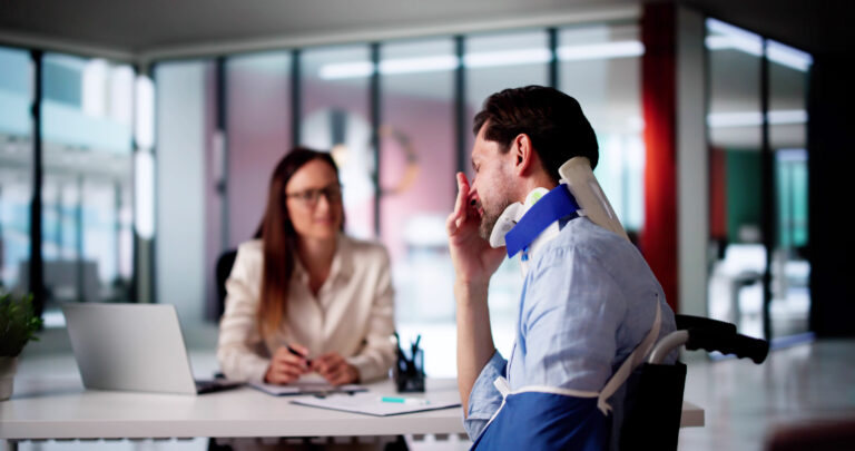 A man wearing a neck brace and arm sling sits at a desk during a consultation, holding his head in discomfort while a professional reviews documents across from him.