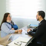 A woman wearing a neck brace and arm sling meets with a professional across a desk, discussing paperwork during a consultation.