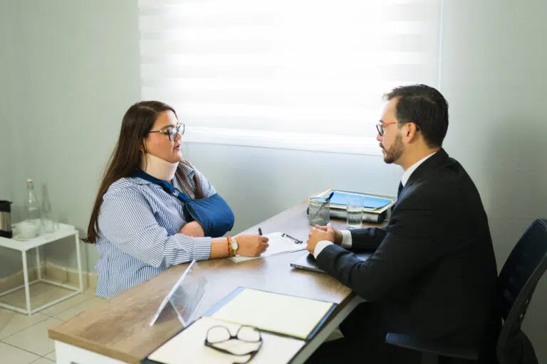 A woman wearing a neck brace and arm sling meets with a professional across a desk, discussing paperwork during a consultation.
