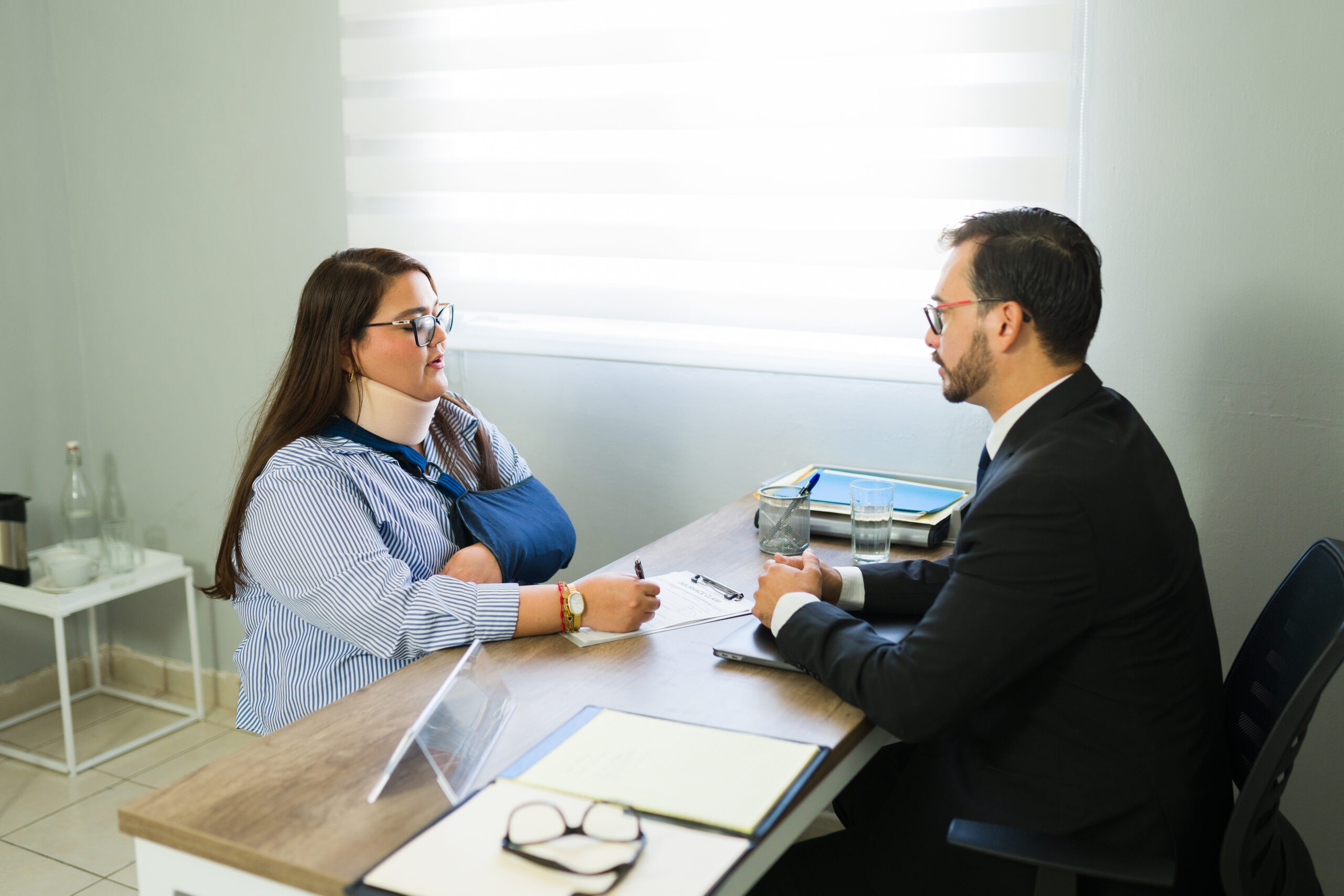 A woman wearing a neck brace and arm sling meets with a professional across a desk, discussing paperwork during a consultation.
