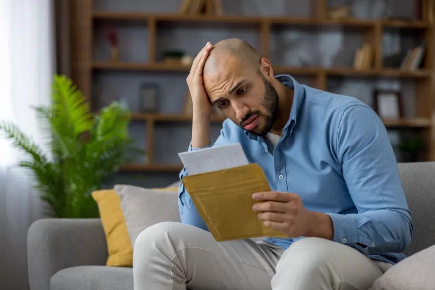 A worried man sits on a couch holding his head while reading a letter, appearing stressed or concerned about its contents.