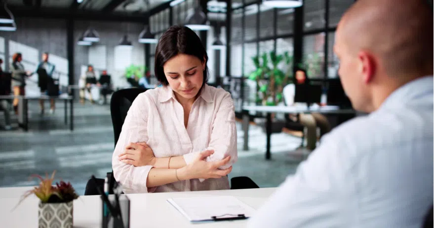 A woman sits at a desk in a modern office with her arms crossed, looking down while speaking with another person across the table. Papers and a clipboard are on the desk.