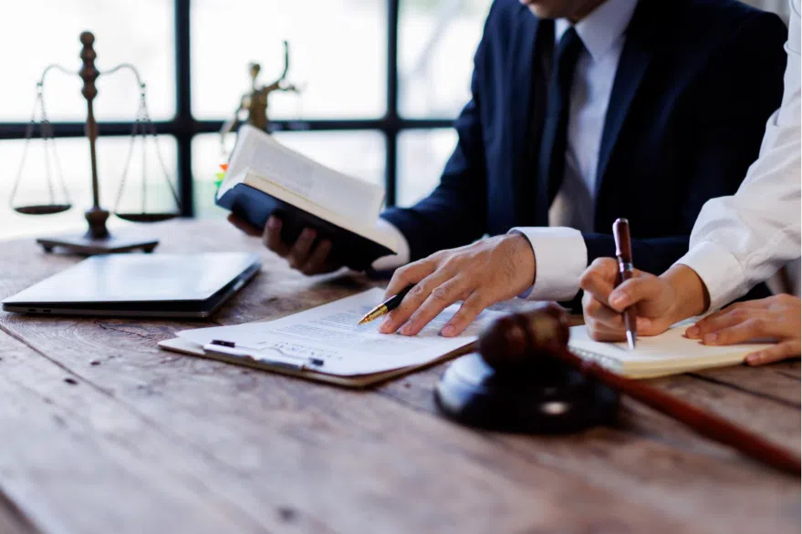 Two attorneys review legal documents at a desk with a gavel and scales of justice nearby, one taking notes while the other reads from a book.