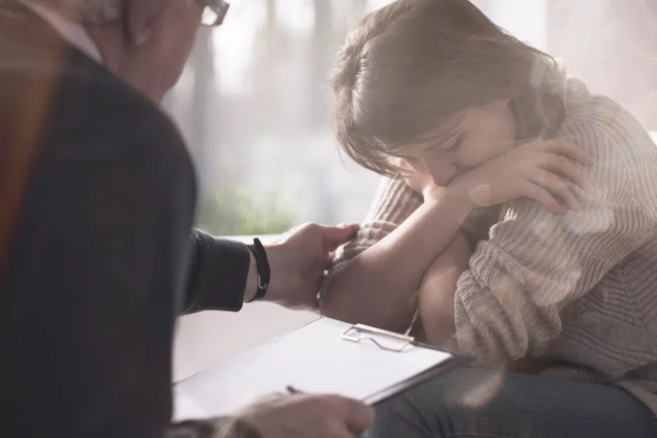 Distressed woman sitting with her head down and arms crossed while a professional seated across from her offers support during a consultation.