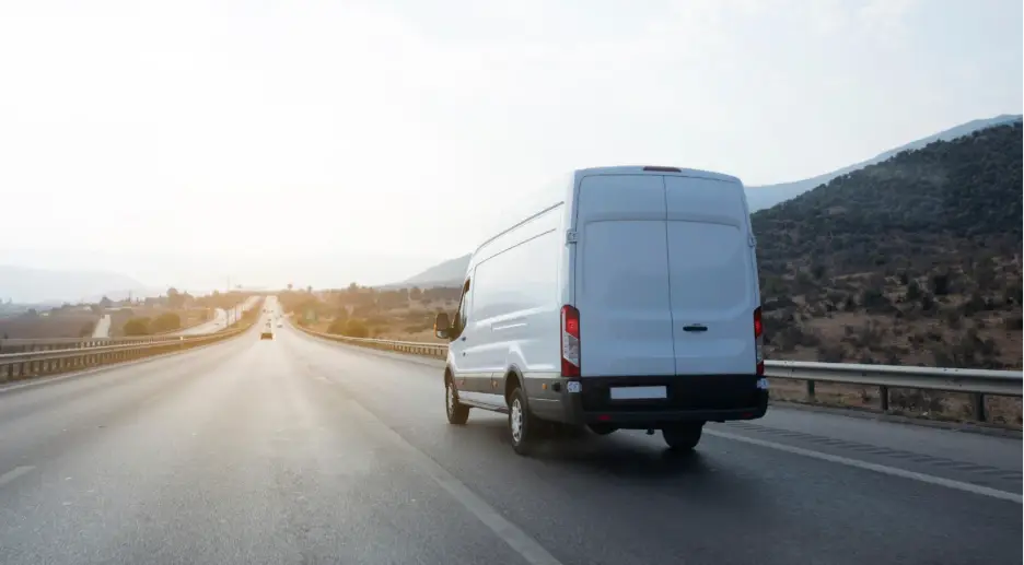 A white cargo van drives along an open highway at sunset, with rolling hills and guardrails lining the road.