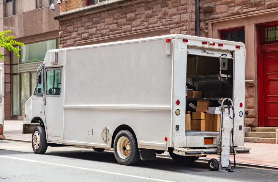 A white box delivery truck is parked on a city street with its rear door open, revealing stacked cardboard boxes and a lowered lift gate.