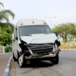 A white delivery van with severe front-end damage is stopped along a roadside, showing a crumpled hood and broken bumper after a collision.