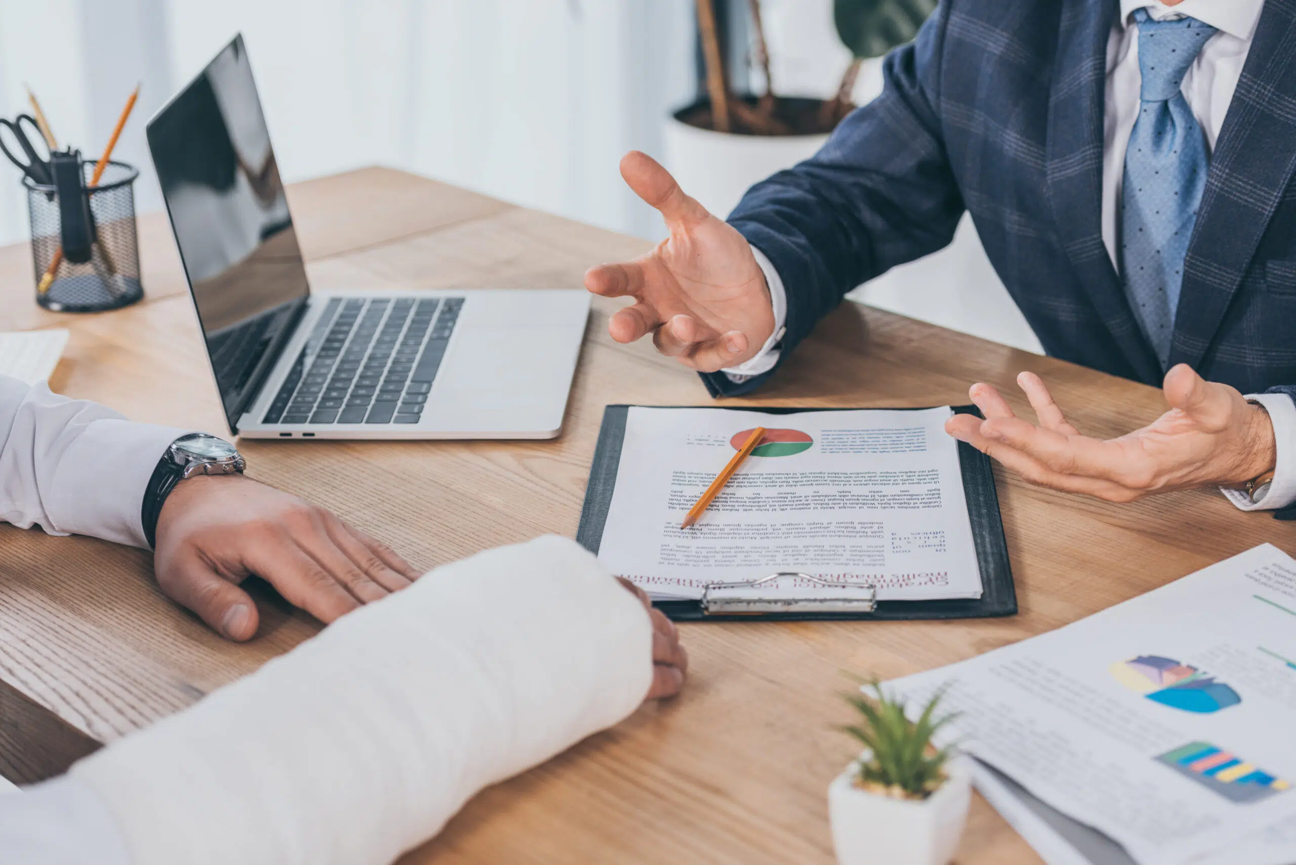 cropped view of businessman in blue jacket siting at table opposite worker with broken arm in office, compensation concept