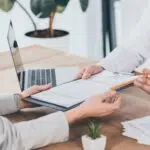 Close-up of two people exchanging a clipboard with documents and a pen across a desk, suggesting signing or reviewing an agreement.