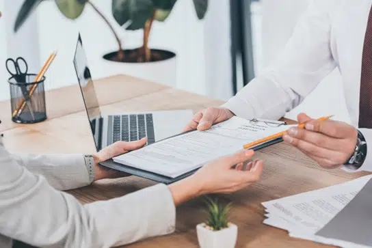 Close-up of two people exchanging a clipboard with documents and a pen across a desk, suggesting signing or reviewing an agreement.