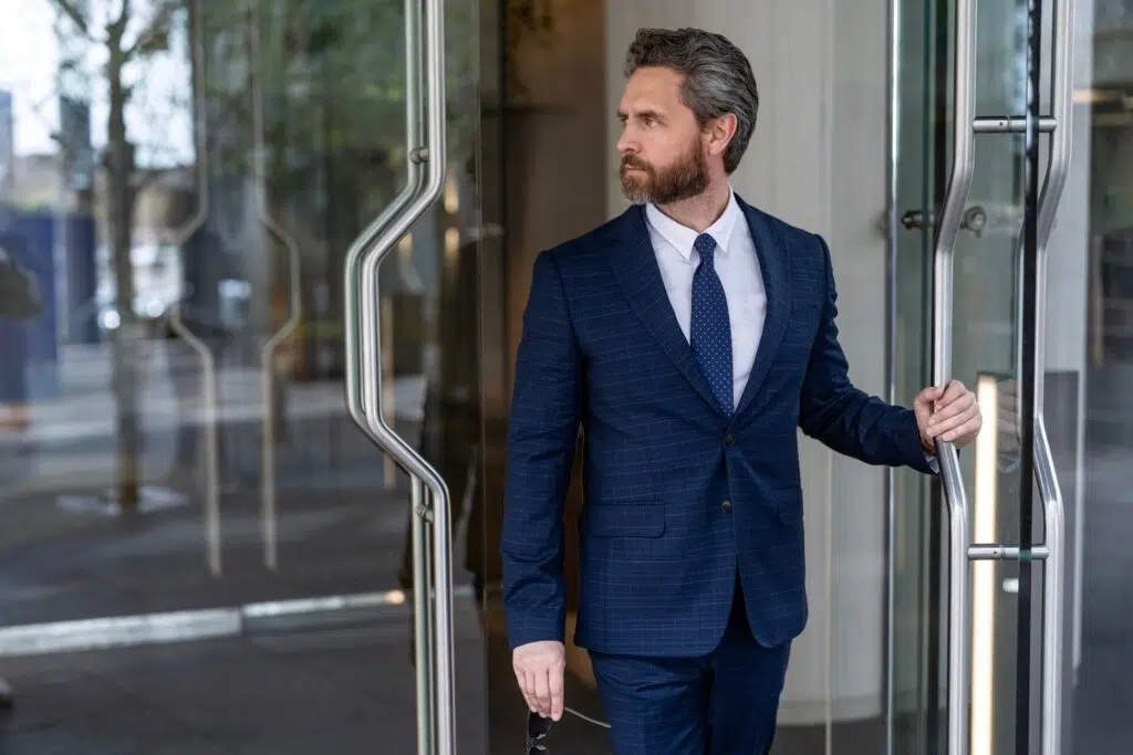 Businessman in a blue suit opening a glass office door while looking outside.