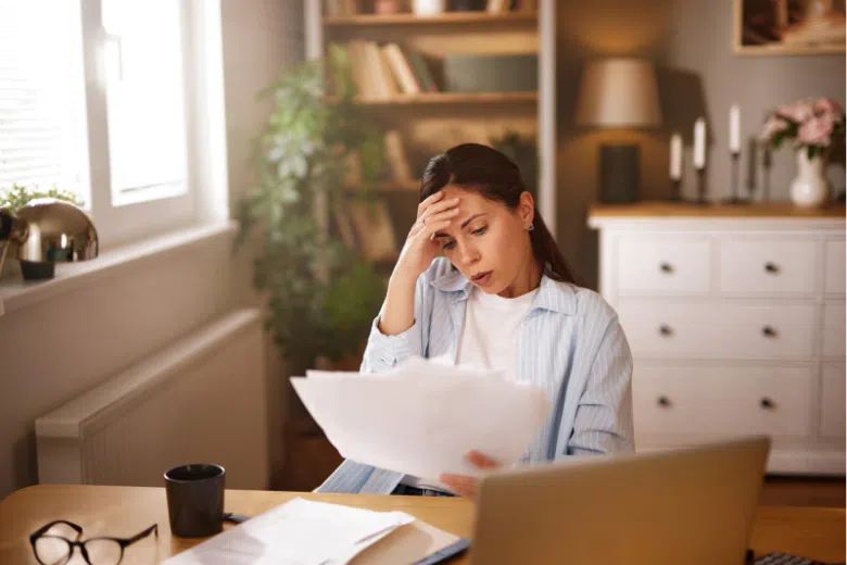 Woman sitting at home looking stressed while reviewing paperwork at a table with a laptop and documents.