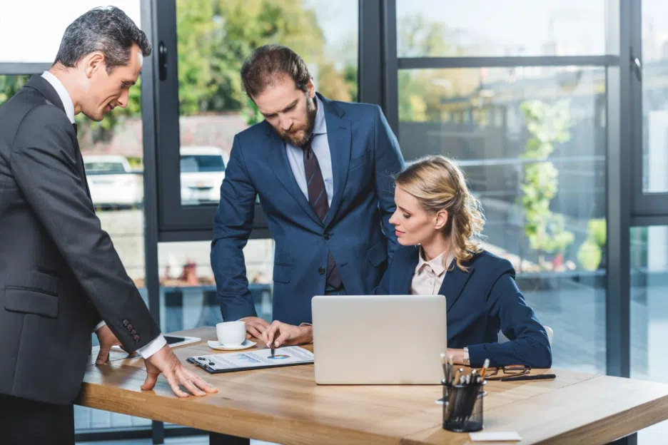 Three business professionals in suits reviewing documents together at a desk with a laptop, collaborating in a modern office setting.
